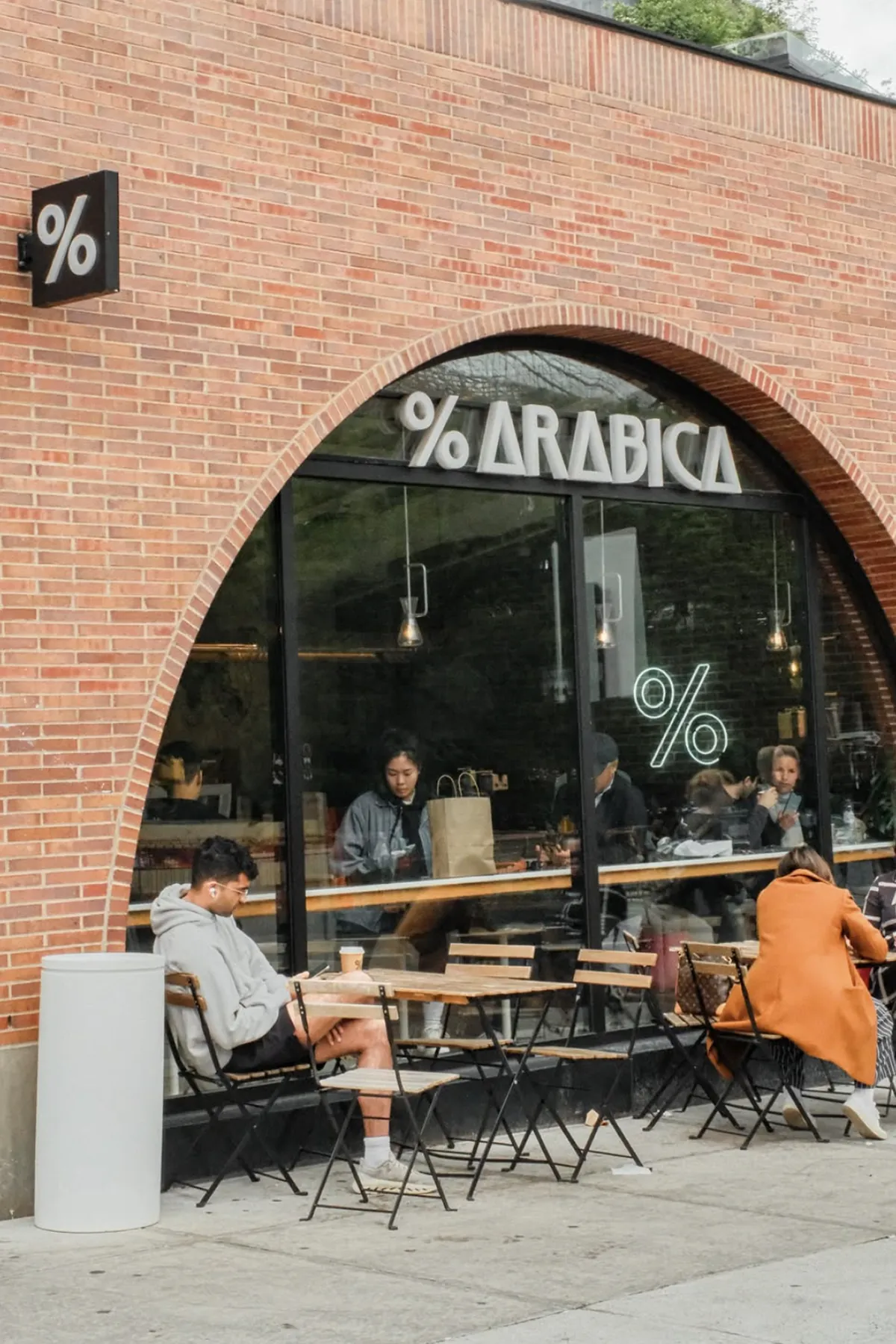 Storefront of % Arabica café featuring a modern brick façade with large arched windows and the % Arabica logo above the entrance. Customers sit at outdoor wooden tables while others enjoy coffee inside, with a glowing % sign visible through the window, capturing the brand’s minimalist design and lively street-side atmosphere.