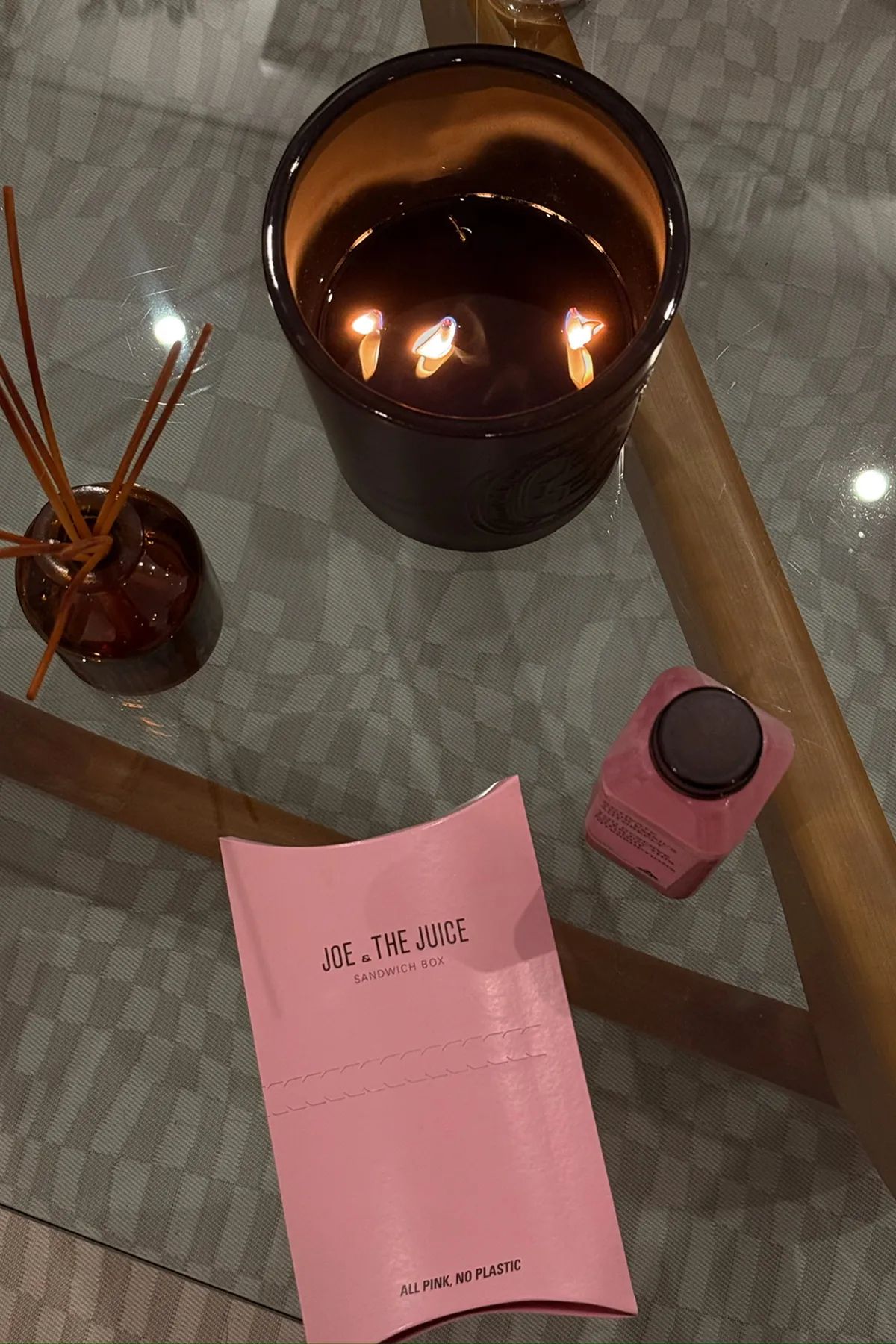 Pink Joe & The Juice sandwich box and a matching pink bottled juice placed on a glass table beside a burning three-wick candle and a small reed diffuser.
