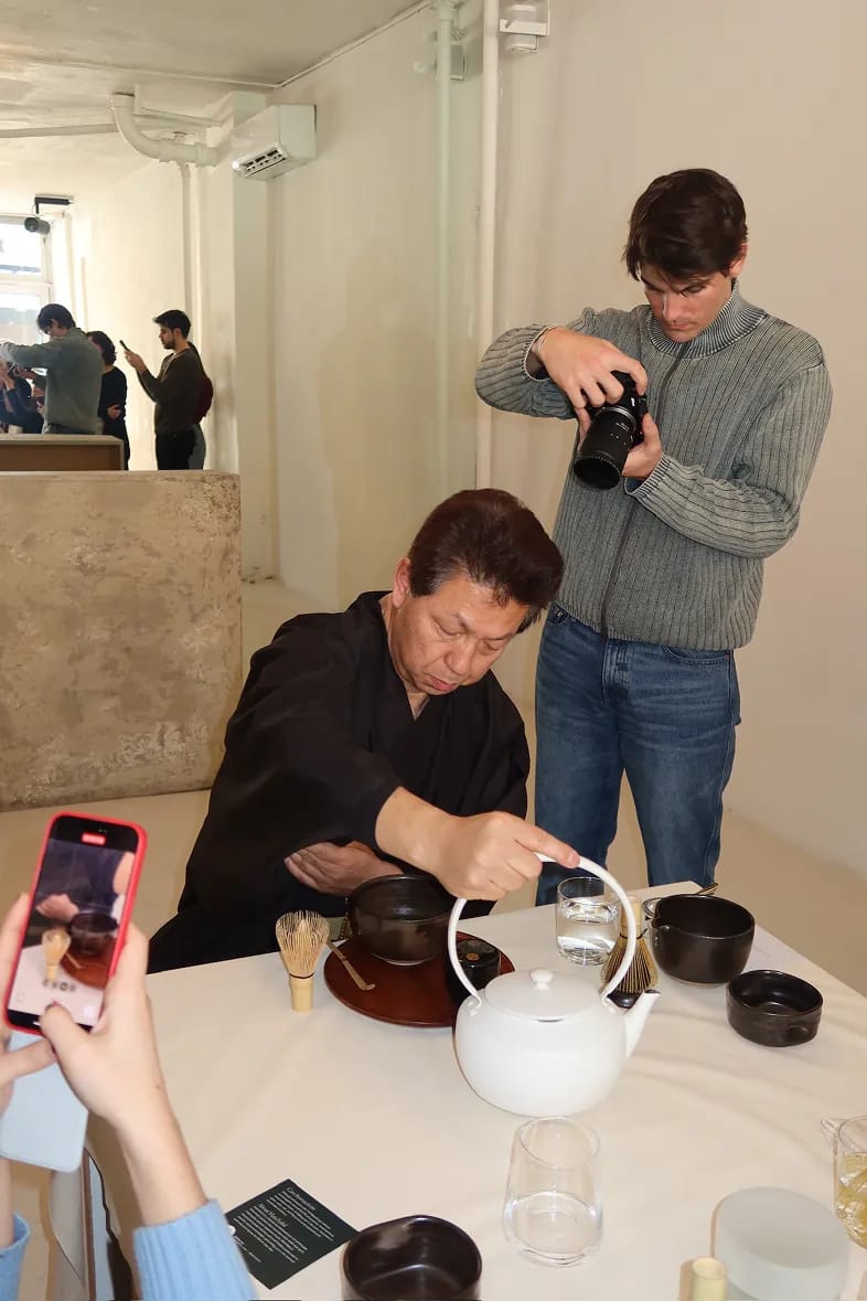 Guests document a traditional matcha tea preparation during a Matchaful x Storytime event in a minimalist gallery space. One attendee prepares tea at a table with ceramic bowls and utensils while others photograph and observe the experience.