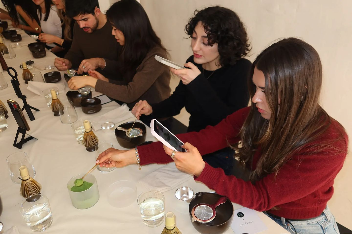 Attendees participate in a hands-on matcha tasting and preparation experience during a Matchaful x Storytime event. Guests sit around a long table using traditional tea tools while photographing and sharing the experience on their phones.