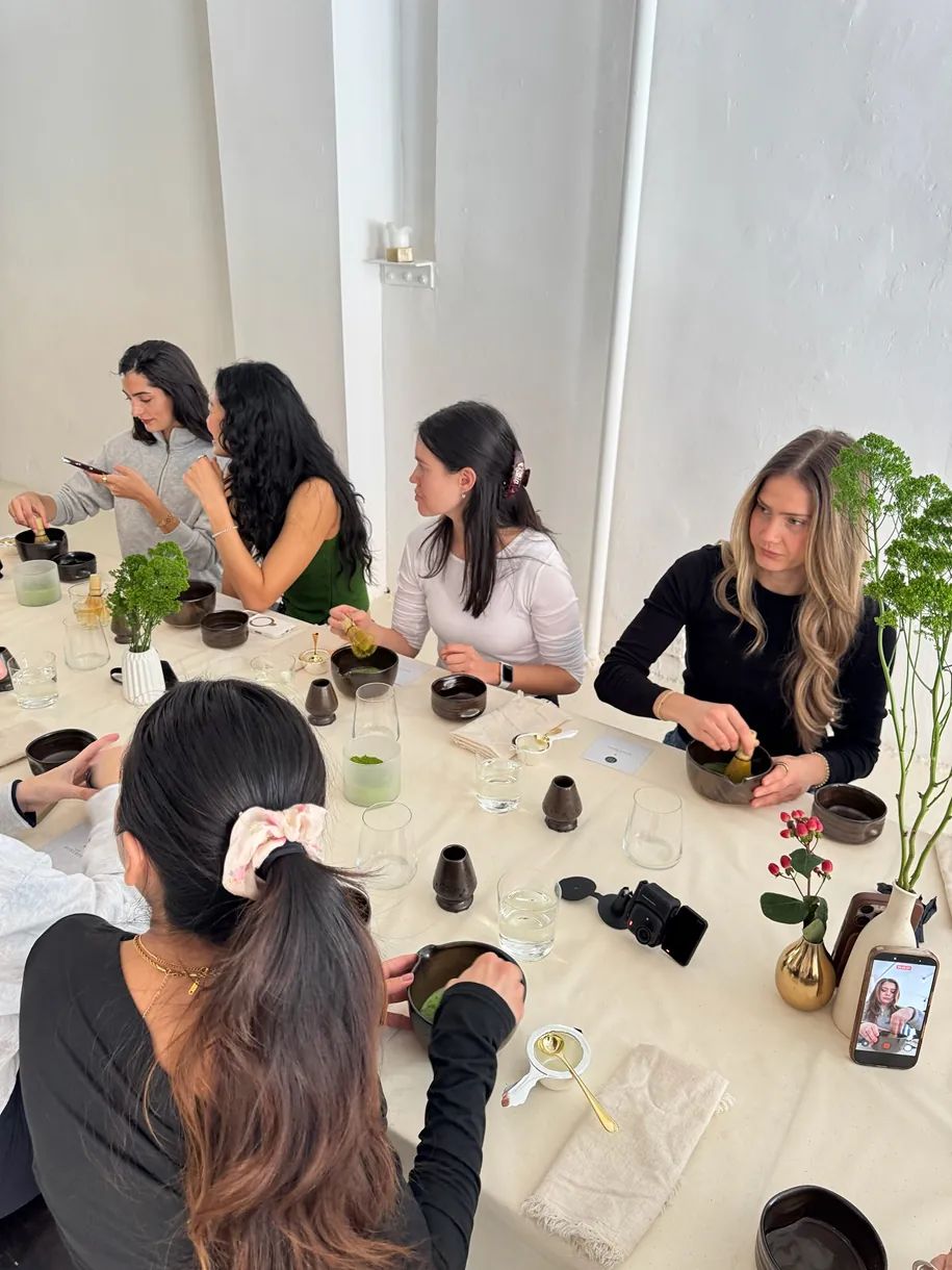 Guests prepare and whisk matcha together during a Matchaful x Storytime event, seated around a decorated communal table with tea bowls, traditional tools, and floral arrangements in a bright studio space.