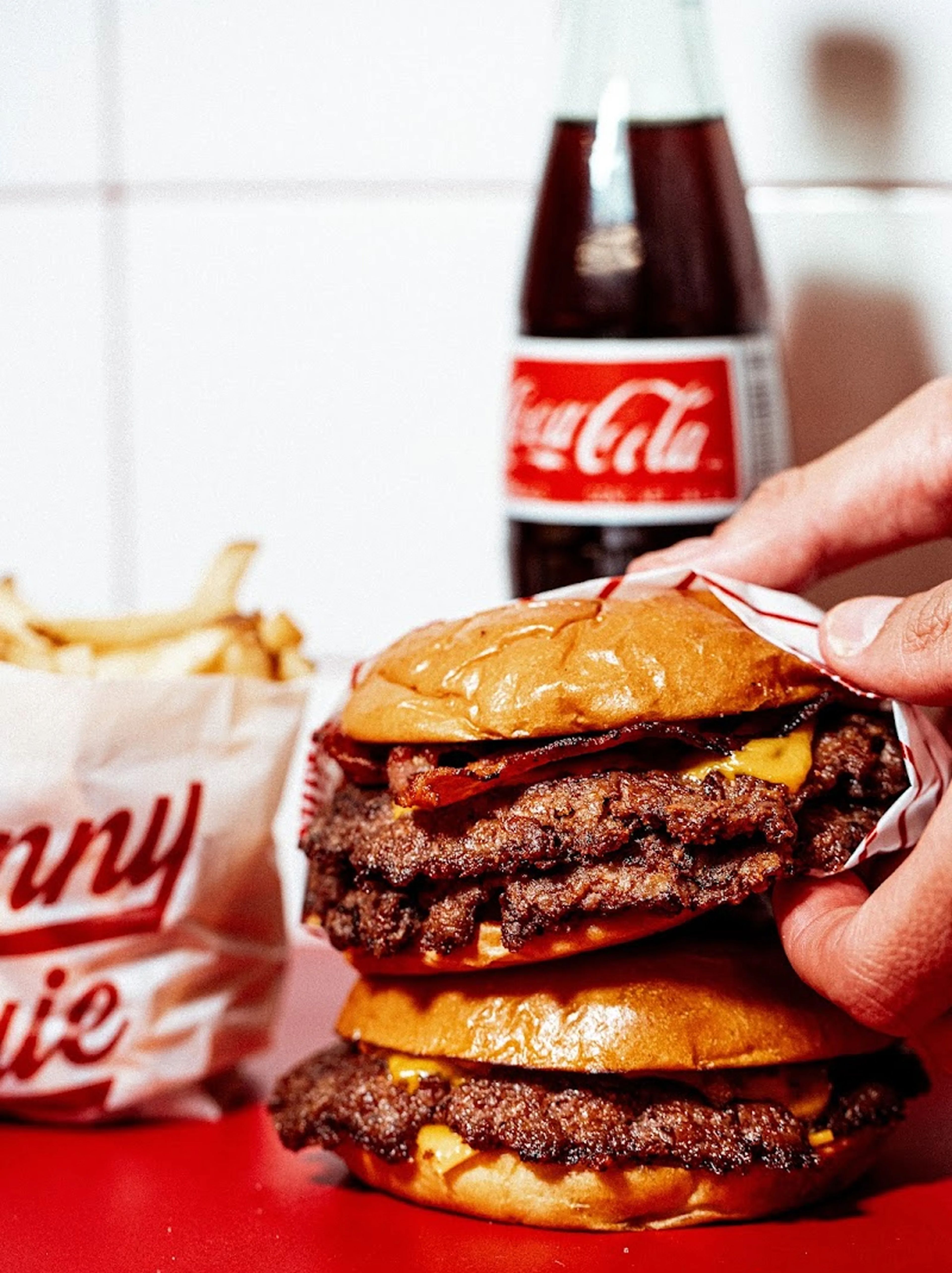 Hand holding a Skinny Louie bacon cheeseburger above another burger, with fries and a Coca-Cola in the background.