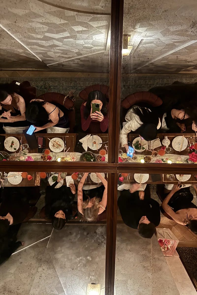 Overhead mirrored view of guests seated at a long dinner table at Beefbar NYC, with candlelight, pink florals, cocktails, and guests capturing the moment on their phones.