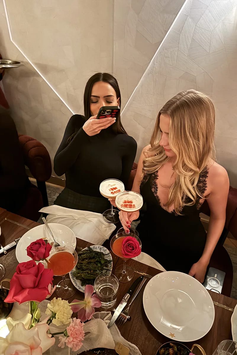@lisaramos and @mollymattporter seated at a candlelit dinner table at Beefbar NYC, holding cocktails garnished with roses, surrounded by pink florals and plated dishes.