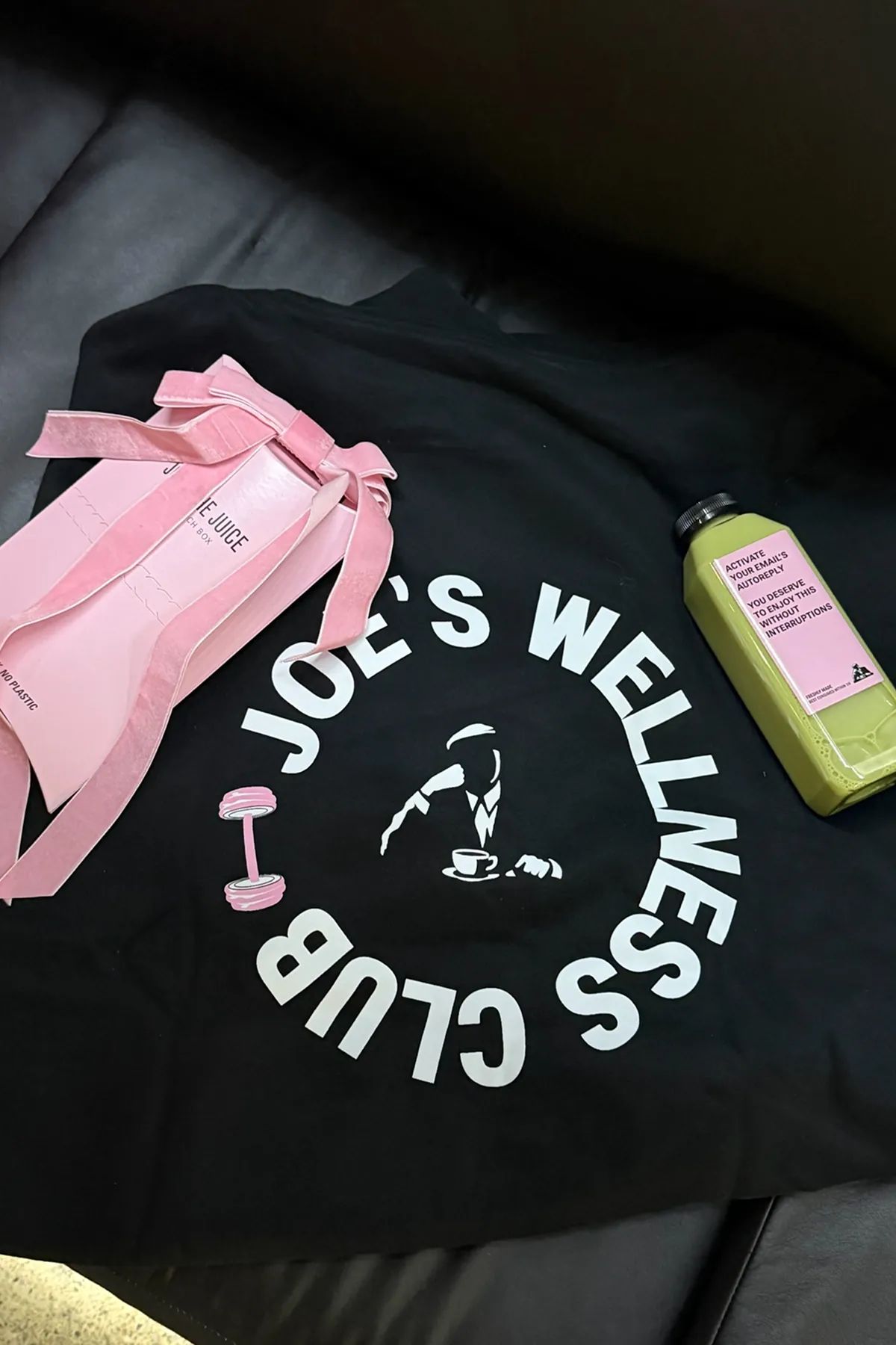 A flat lay featuring a Joe & The Juice Wellness Club hoodie, a pink Joe and The Juice sandwich box tied with a velvet bow, and a green bottled juice with a pink Joe and The Juice label, displayed on a black surface.
