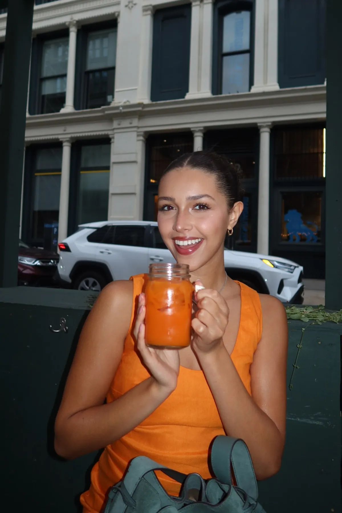 @ms.joynavarro at the Le Botaniste x Storytime dinner holds a bright orange botanical drink in a mason jar while seated outdoors. The scene includes a backdrop of city buildings and a relaxed, friendly atmosphere.