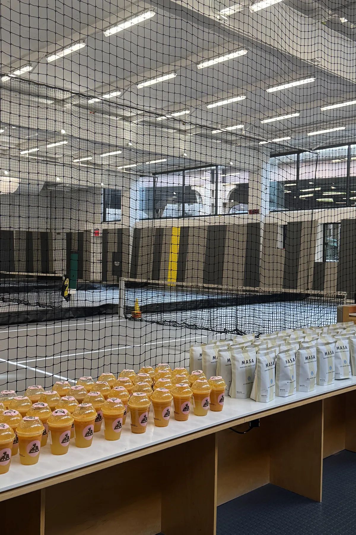 Joe and The Juice smoothies and MASA tortilla chips bags arranged on a table at Court 16, with indoor tennis courts visible behind a safety net in a bright, modern facility.