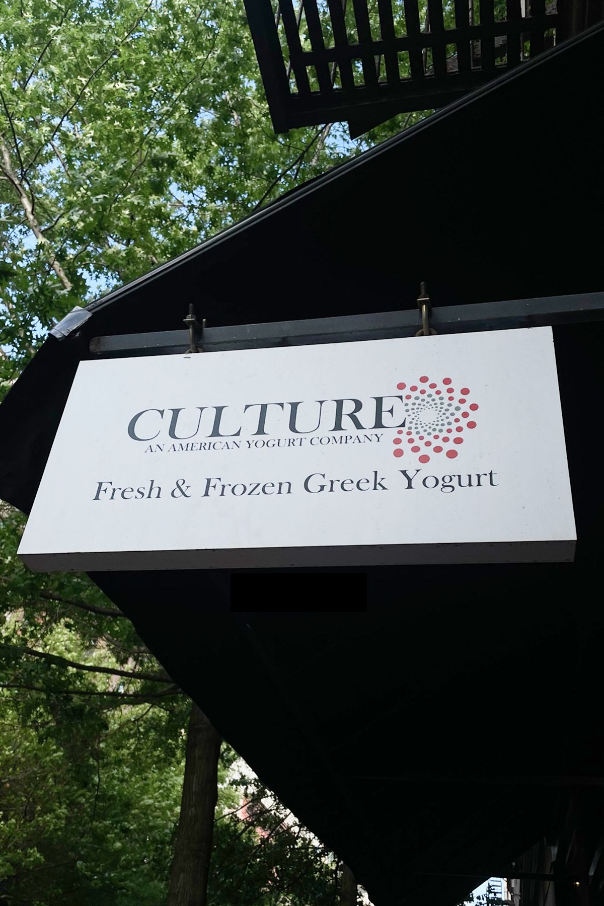 Storefront sign for Culture, An American Yogurt Company, displaying the tagline “Fresh & Frozen Greek Yogurt.” The sign hangs under a black awning with green trees visible in the background, highlighting the shop’s outdoor street presence.