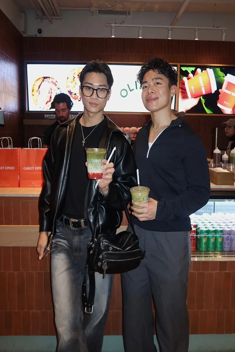 Two creators in black leather and navy pose with Carrot Express juices at the counter at Carrot Express
