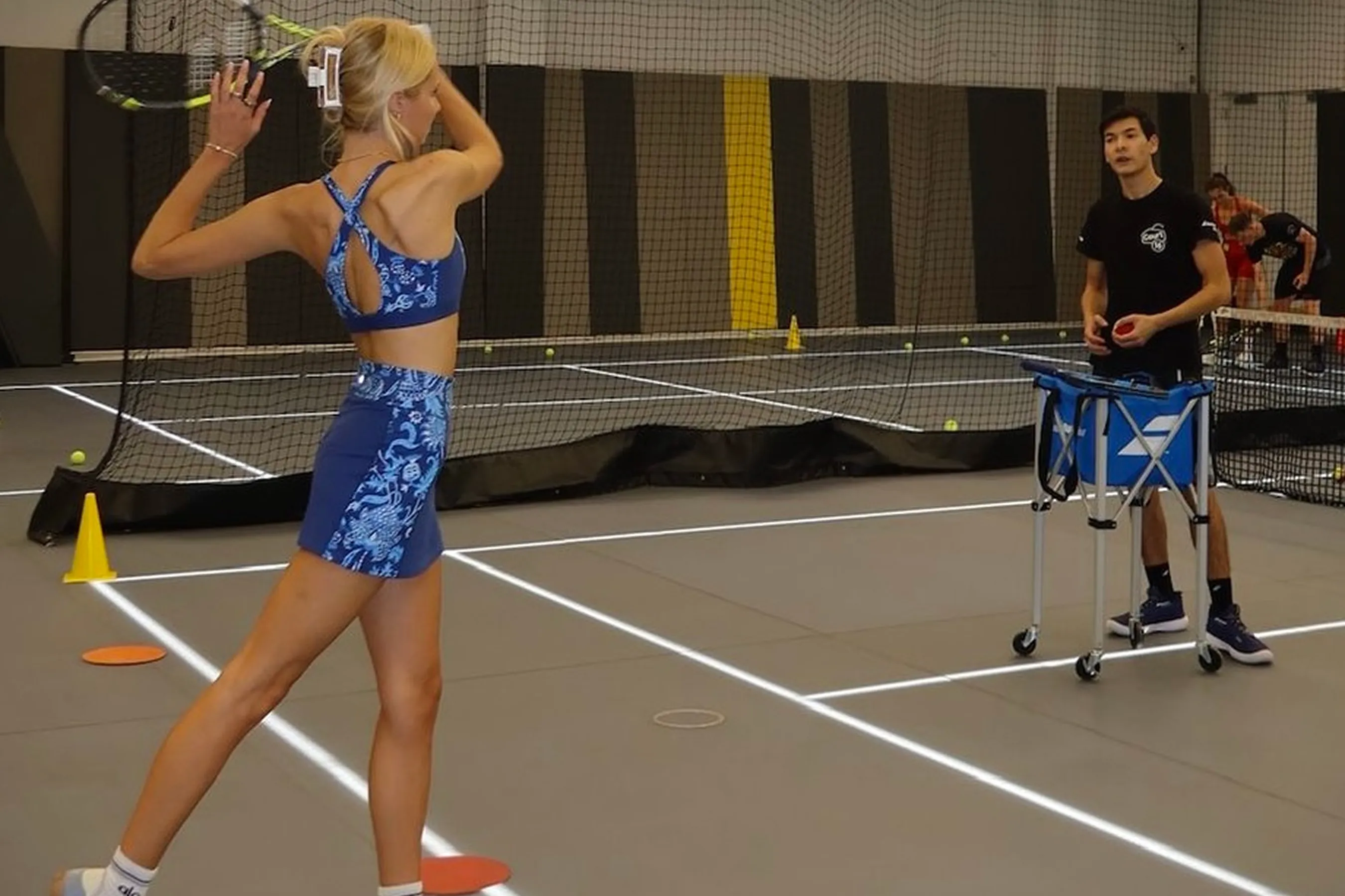 @abbygendell wearing a blue athletic set practices her tennis swing during a Court 16 session, while an instructor stands nearby preparing tennis balls. The indoor court is marked with training cones and surrounded by safety nets.