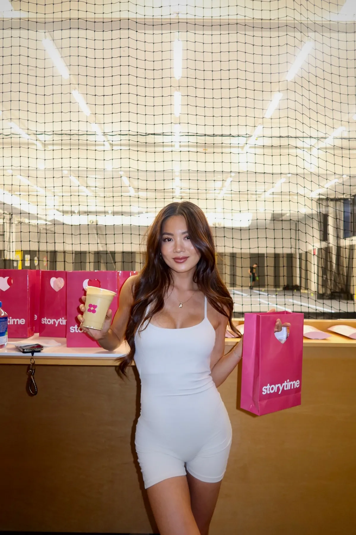 Storytime influencer wearing a white athletic romper poses at a Court 16 event, holding a Brasa Peruvian Kitchen smoothie in one hand and a pink Storytime goodie bag in the other. Behind her, more Storytime bags are displayed in front of the indoor tennis courts.