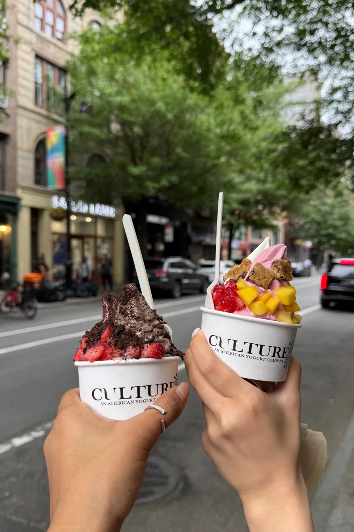 Two hands hold cups of Culture An American Yogurt Company frozen yogurt on a New York City street. One cup features chocolate soft serve topped with strawberries and crushed Oreos, while the other has vanilla and pink swirl froyo topped with mango, strawberries, and cookie chunks. The background shows a busy urban street with cars, storefronts, and trees, capturing the fresh, colorful, and photogenic appeal of Culture’s popular frozen yogurt.