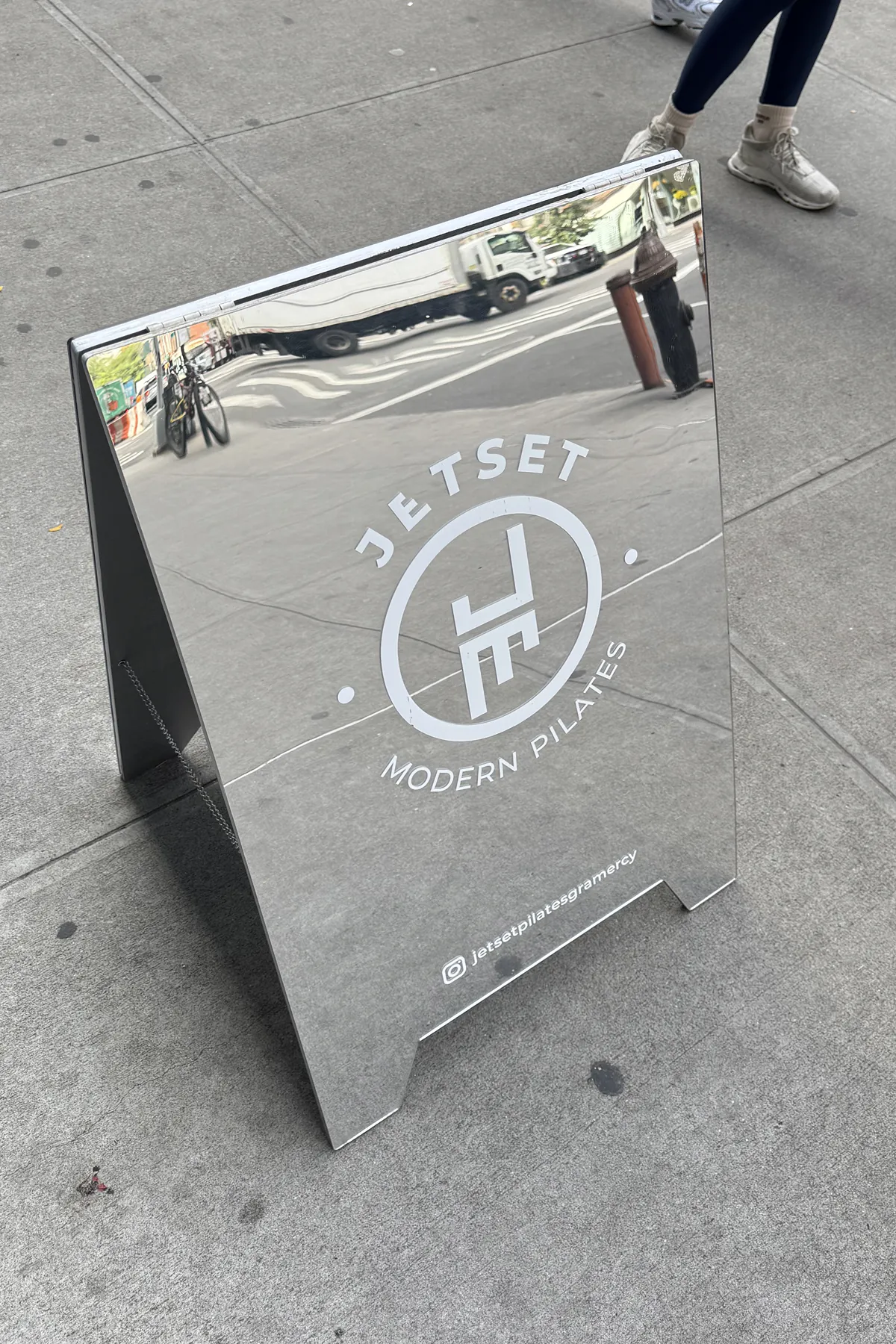 A reflective JetSet Modern Pilates sidewalk sign displayed outside the studio entrance in NYC, showing the JetSet logo and Instagram handle against the city street backdrop.