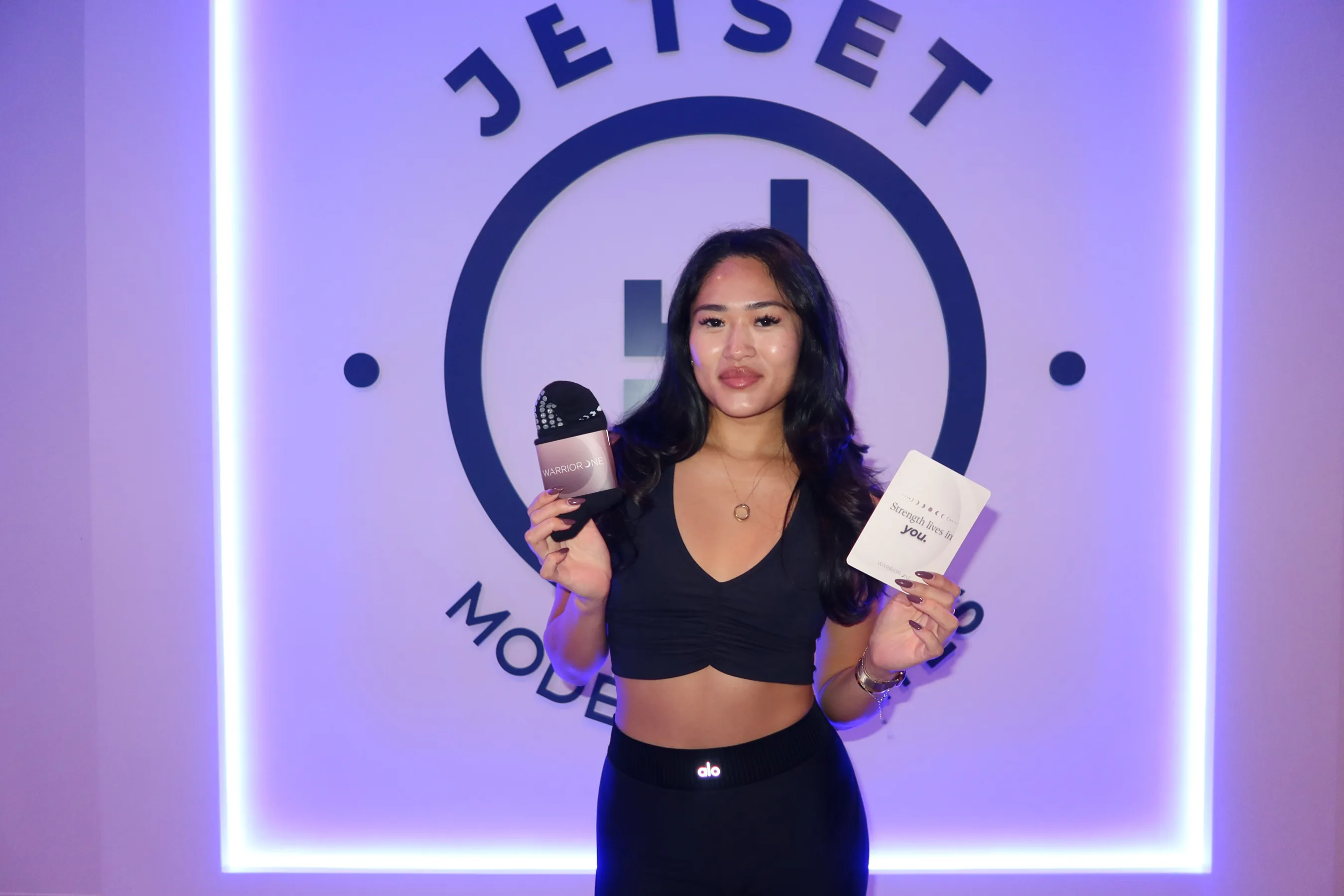 @paveekwun standing in front of a glowing JetSet Pilates logo wall, holding a pair of @warrioroneofficial grip pilates socks in one hand and a small inspirational card in the other. She is wearing a black workout outfit and smiling softly at the camera under purple-blue lighting.