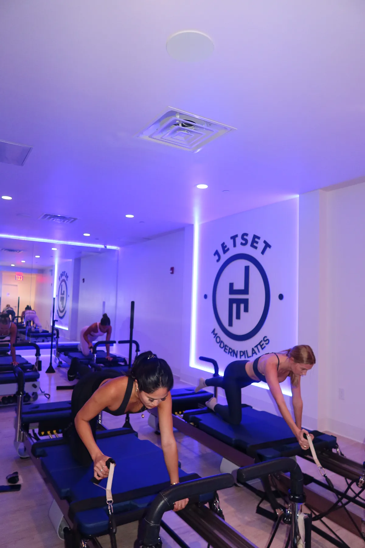 @paveekwun and @mollymattporter taking a high-intensity Pilates class at JetSet Pilates, wearing Alo Yoga, using reformer machines in a studio lit with soft blue and purple lighting. Two women are in the foreground balancing in a plank-like position while holding straps, with the JetSet Modern Pilates logo glowing on the wall behind them.