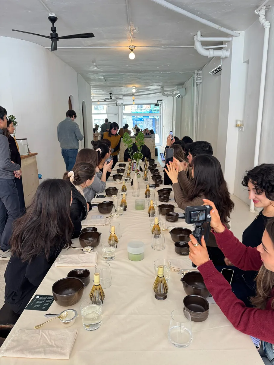 Storytime creators gather around a long communal table during a Matchaful workshop, documenting the experience while learning traditional matcha preparation in a minimalist NYC studio.
